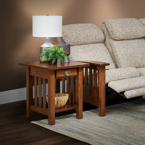 Mission oak end table with drawer, vertical slats and lower shelf holding a basket beside a reclining sofa