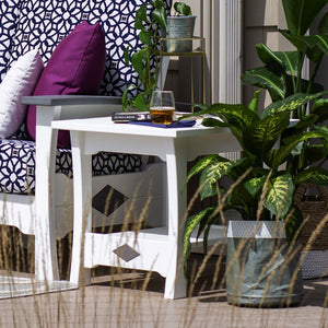 White poly patio end table with diamond cutout accents and a drink and book on top beside tropical plants