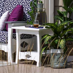 White poly patio end table with diamond cutout accents and a drink and book on top beside tropical plants