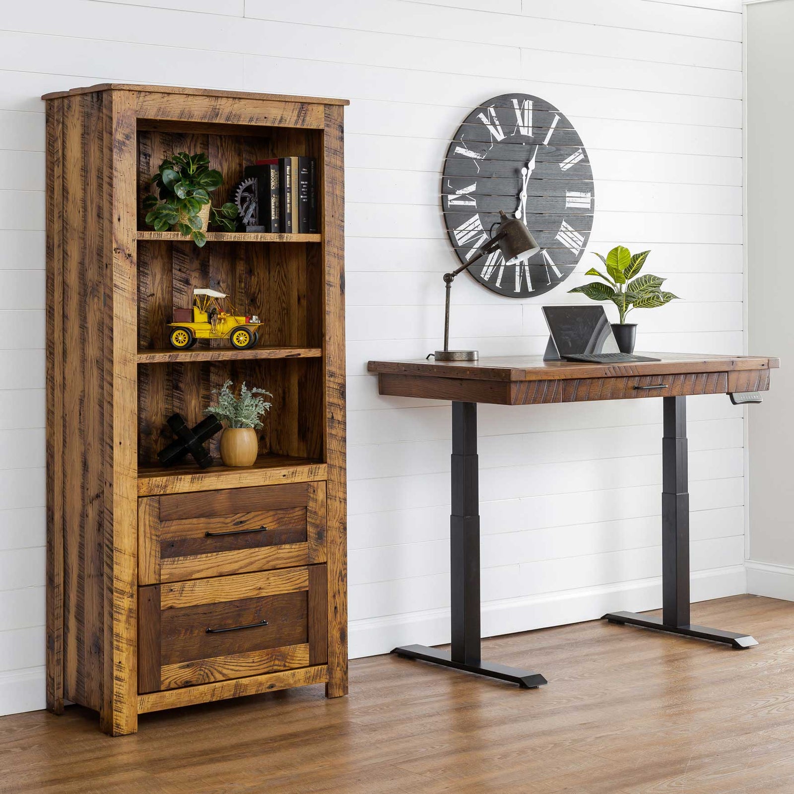 Reclaimed wood bookcase with two file drawers styled with books and plants beside a black-base standing desk and large wall clock