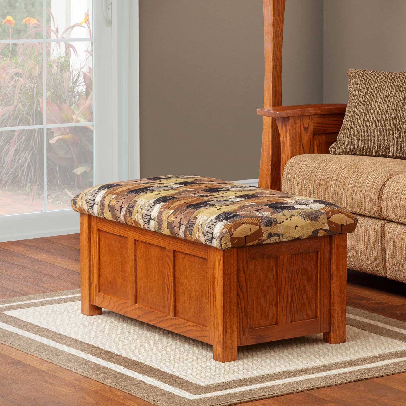 Oak cushion storage ottoman with raised panel sides and patterned upholstered lid beside a sofa by a window