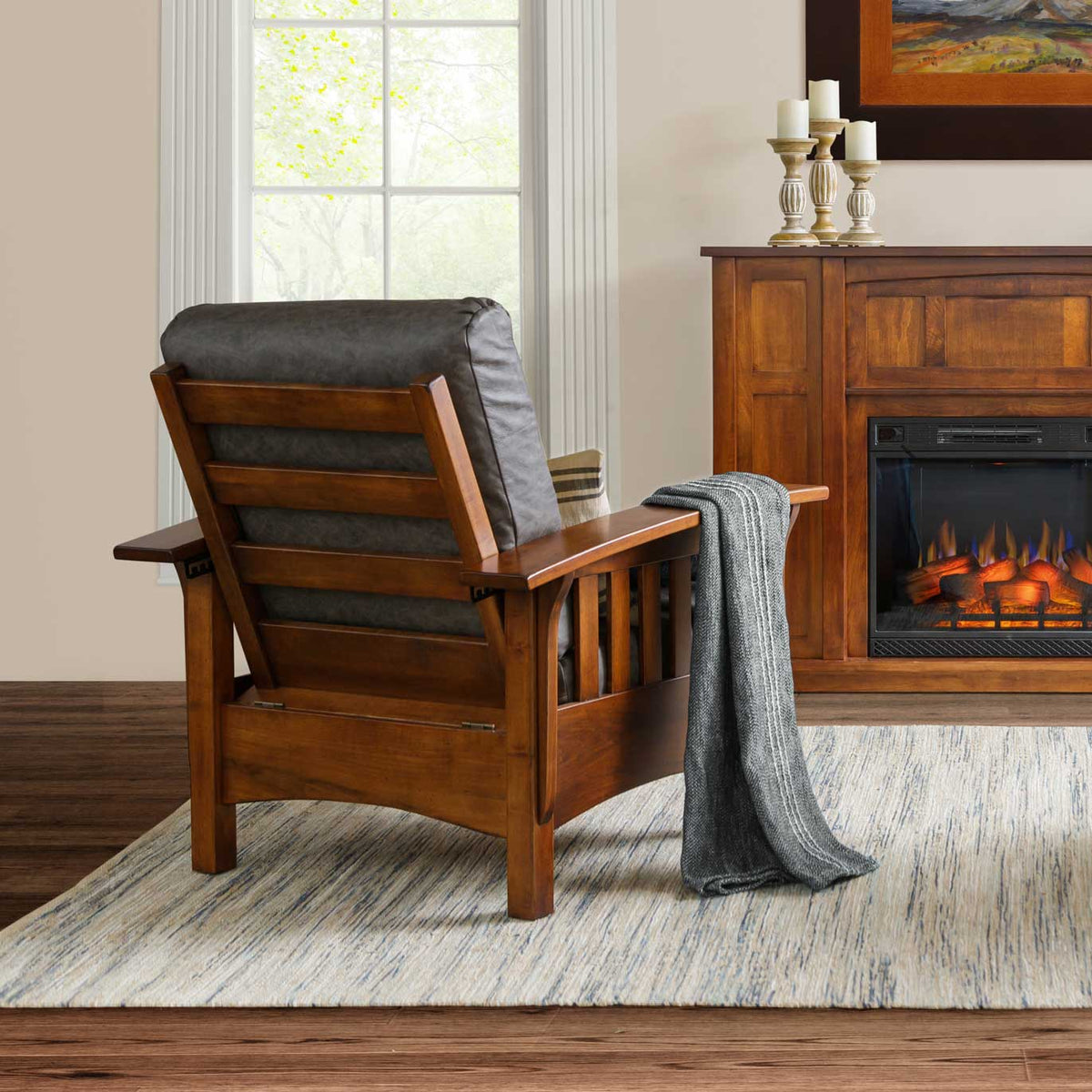Rear view of a cherry Morris chair with gray leather cushions and a throw, beside an electric fireplace