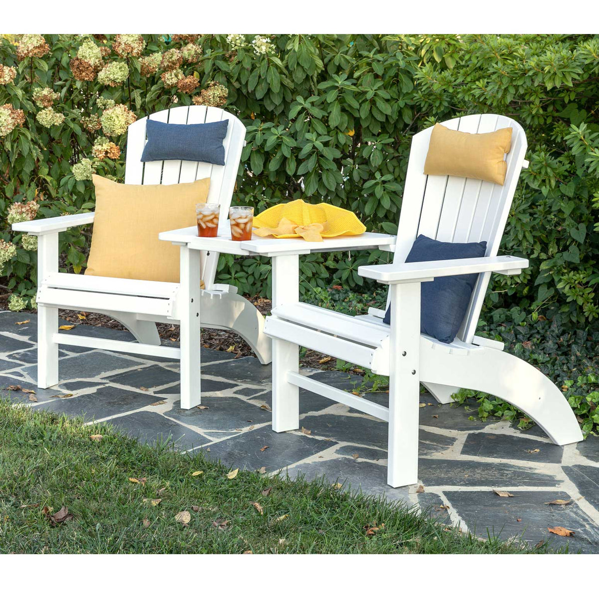 Two white fanback Adirondack chairs linked by a built-in poly side table with pillows and iced tea on a slate patio