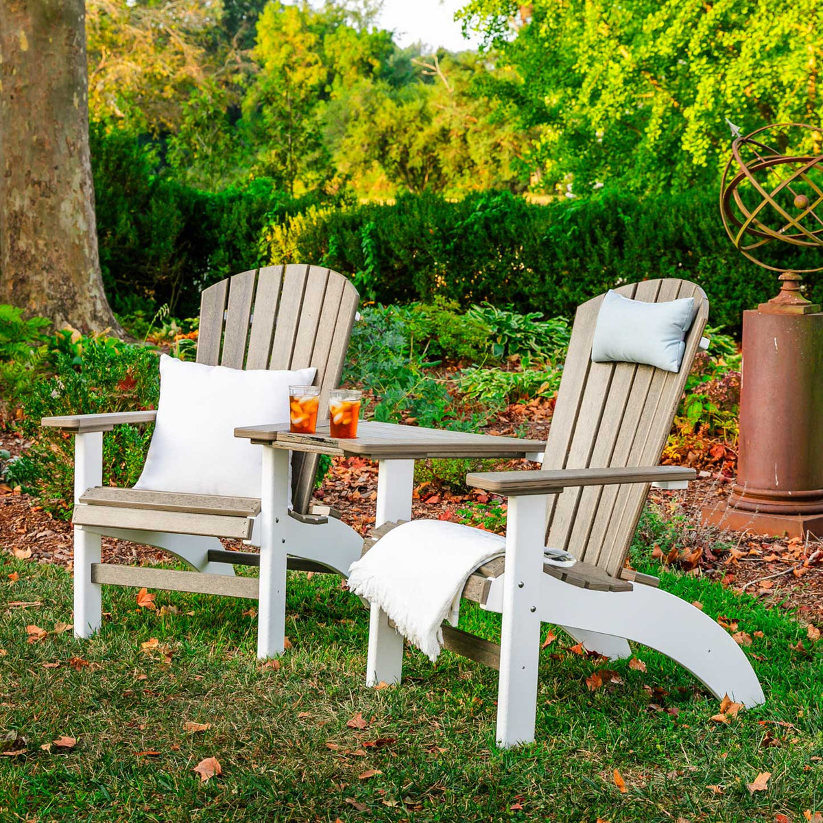 Pair of weathered gray fanback Adirondack chairs with white bases joined by a poly tete-a-tete table in a garden setting