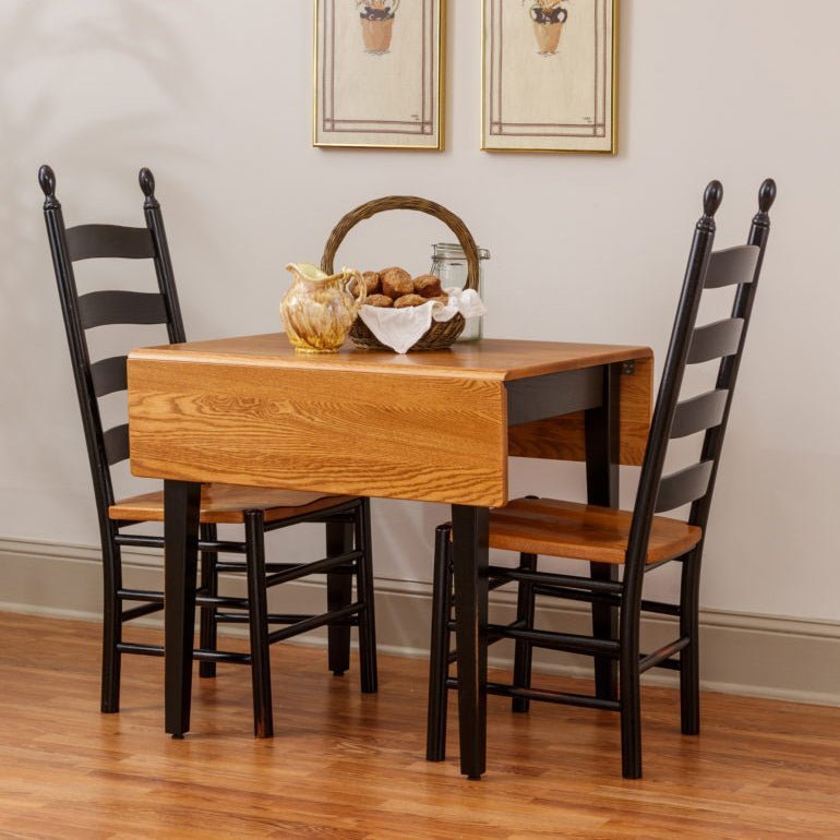 Small oak drop leaf dining table with one leaf down and two tall black ladder back chairs with wood seats