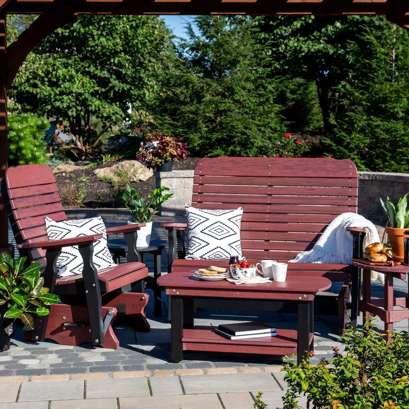Cherrywood and black poly glider chair and double glider with an oval coffee table under a wood pergola on a stone patio