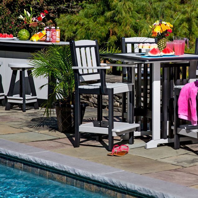 Black and white poly bar height patio chairs around a tall square table beside a swimming pool with fruit and drinks