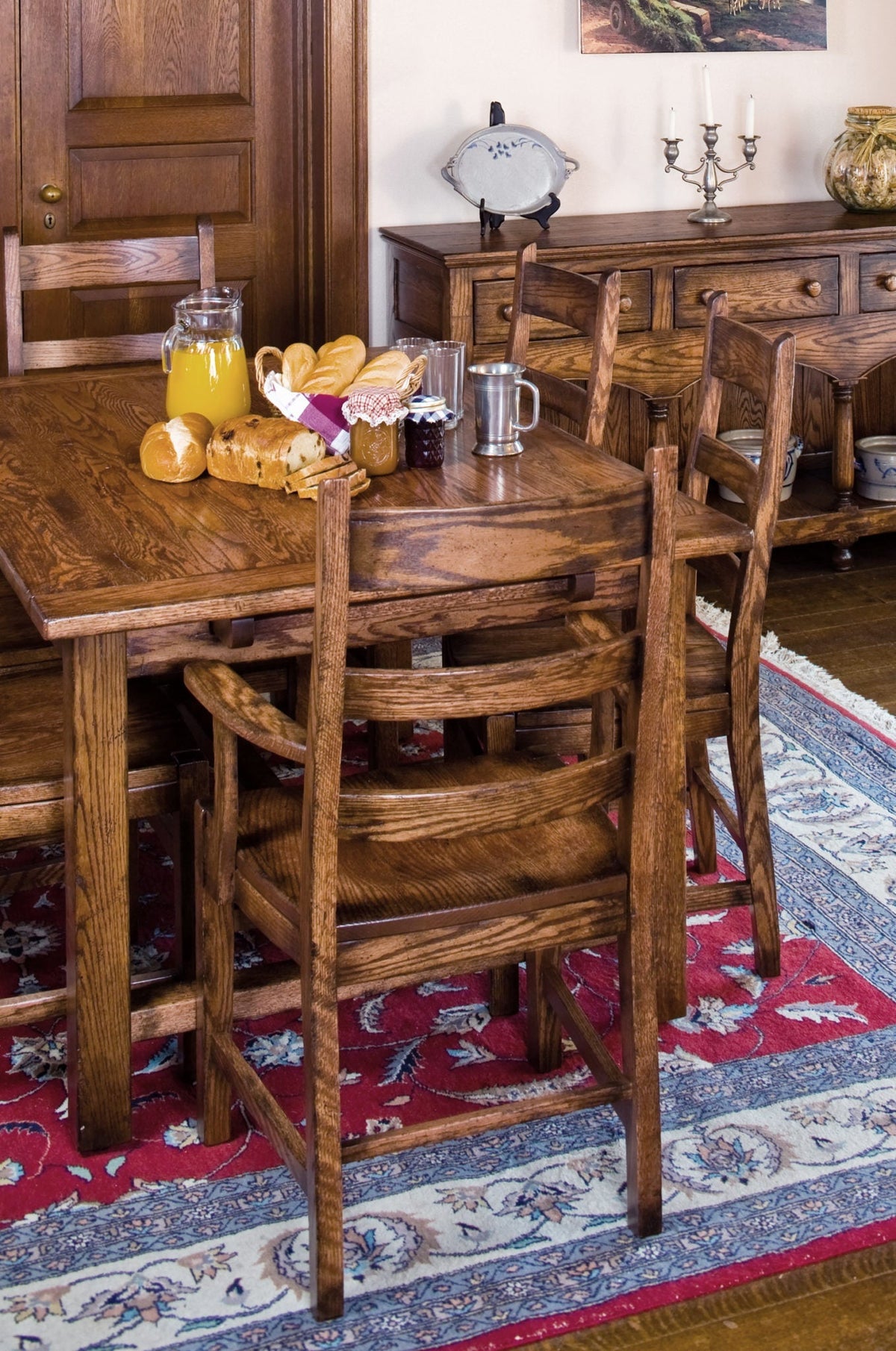 Oak ladder back chairs with wood seats at a farmhouse table set with bread and juice on a red patterned rug