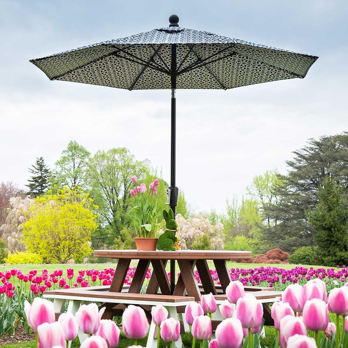 Black and tan patterned patio umbrella shading a brown poly picnic table in a garden of pink tulips
