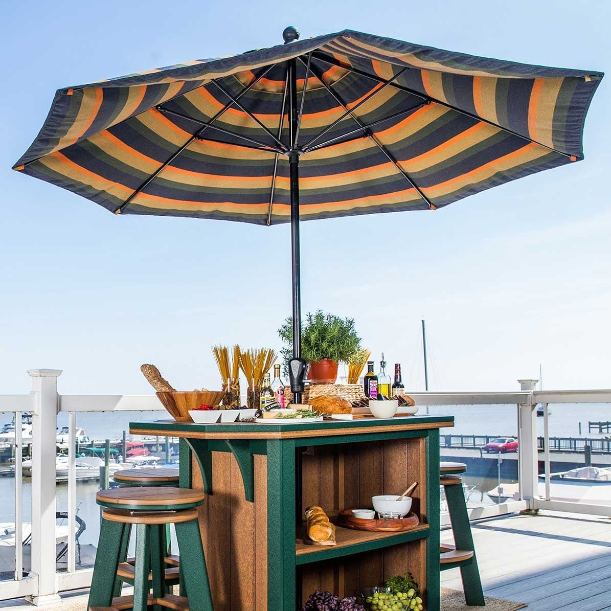 Striped navy and orange market umbrella over a green and cedar poly bar table with swivel stools at a marina