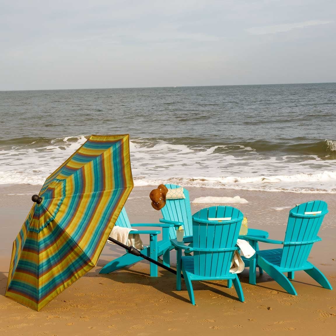 Tilted teal and gold striped umbrella beside turquoise poly Adirondack chairs on a sandy beach