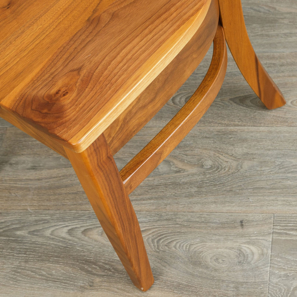 Detail of the walnut dining chair's scooped seat edge, curved side stretcher and gently splayed rear leg on gray plank flooring