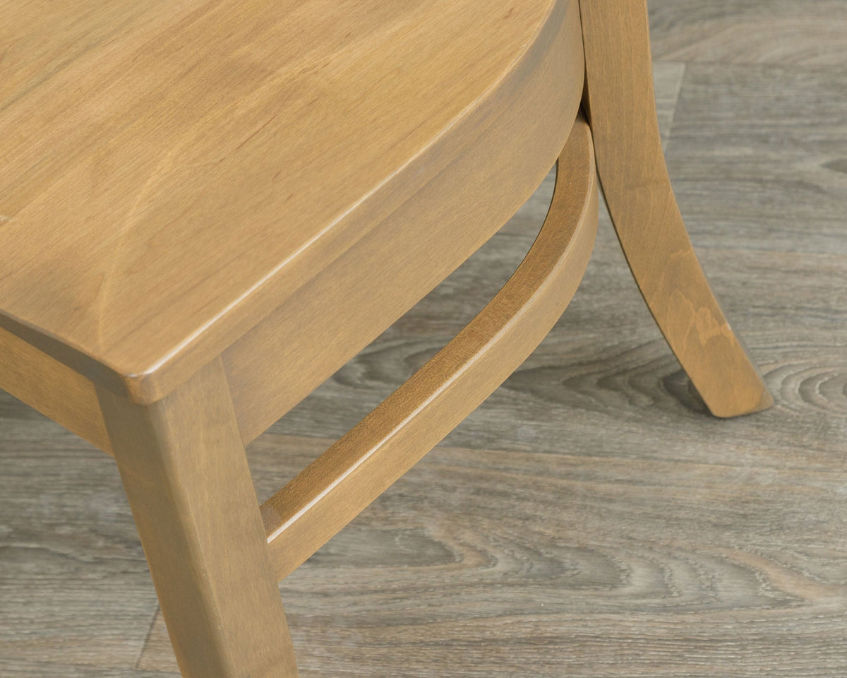 Detail of the light finish dining chair's solid seat corner, apron and side stretchers on gray wood flooring