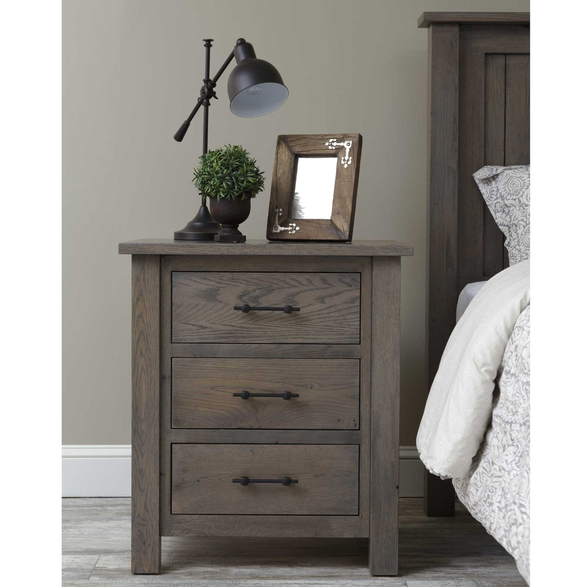 Rustic gray oak nightstand with three drawers styled with a black task lamp, plant and picture frame beside a bed