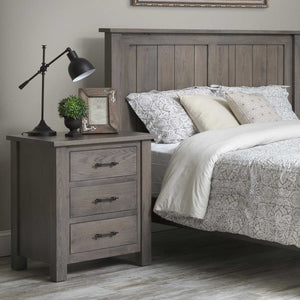 Weathered gray three drawer nightstand with black bar pulls beside a matching plank panel bed and reading lamp