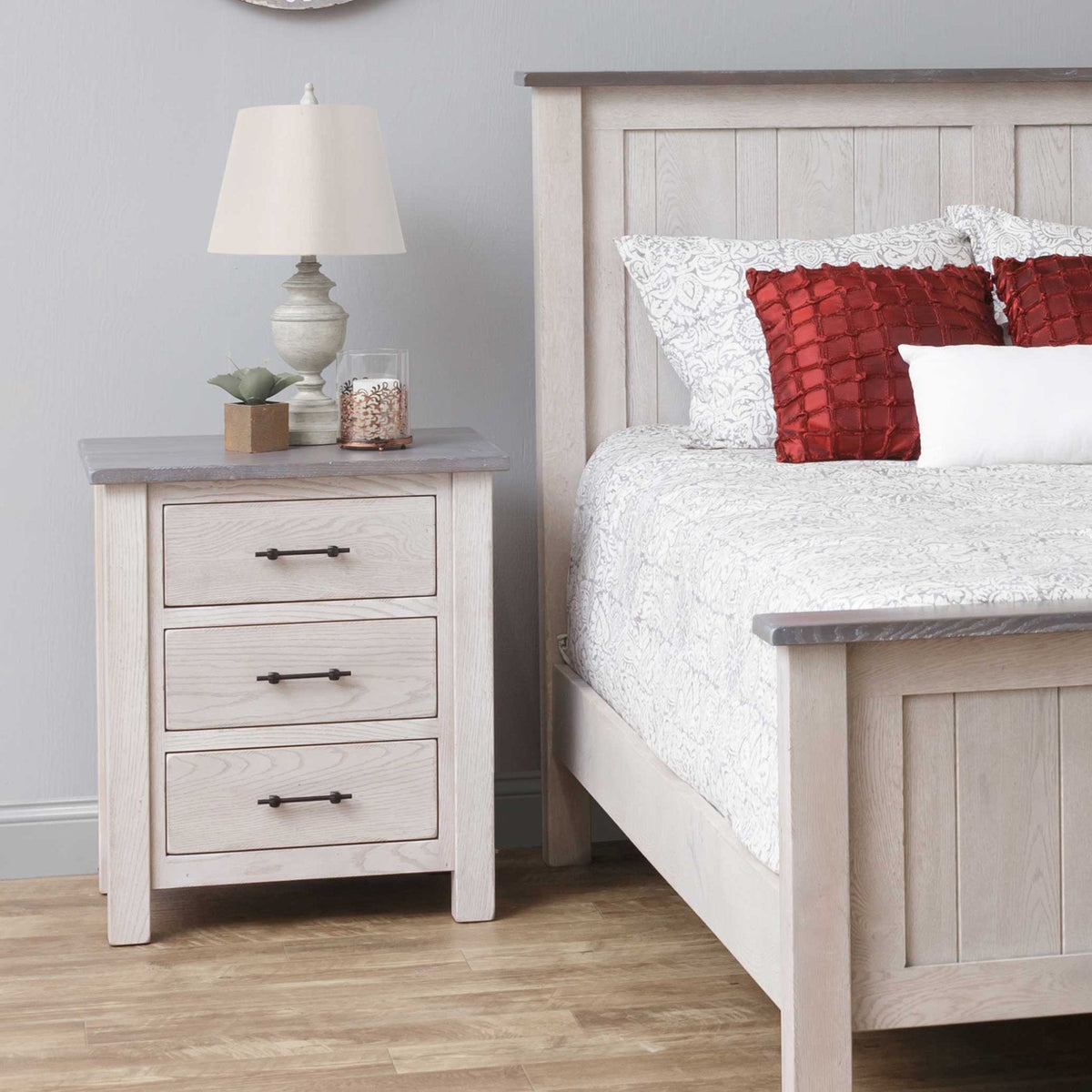 Whitewashed oak three drawer nightstand with gray top and black pulls, staged next to a matching panel bed with red pillows