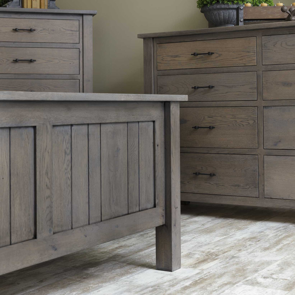 Foot of the gray plank bed with the eight drawer dresser and chest of drawers behind on light wood floors
