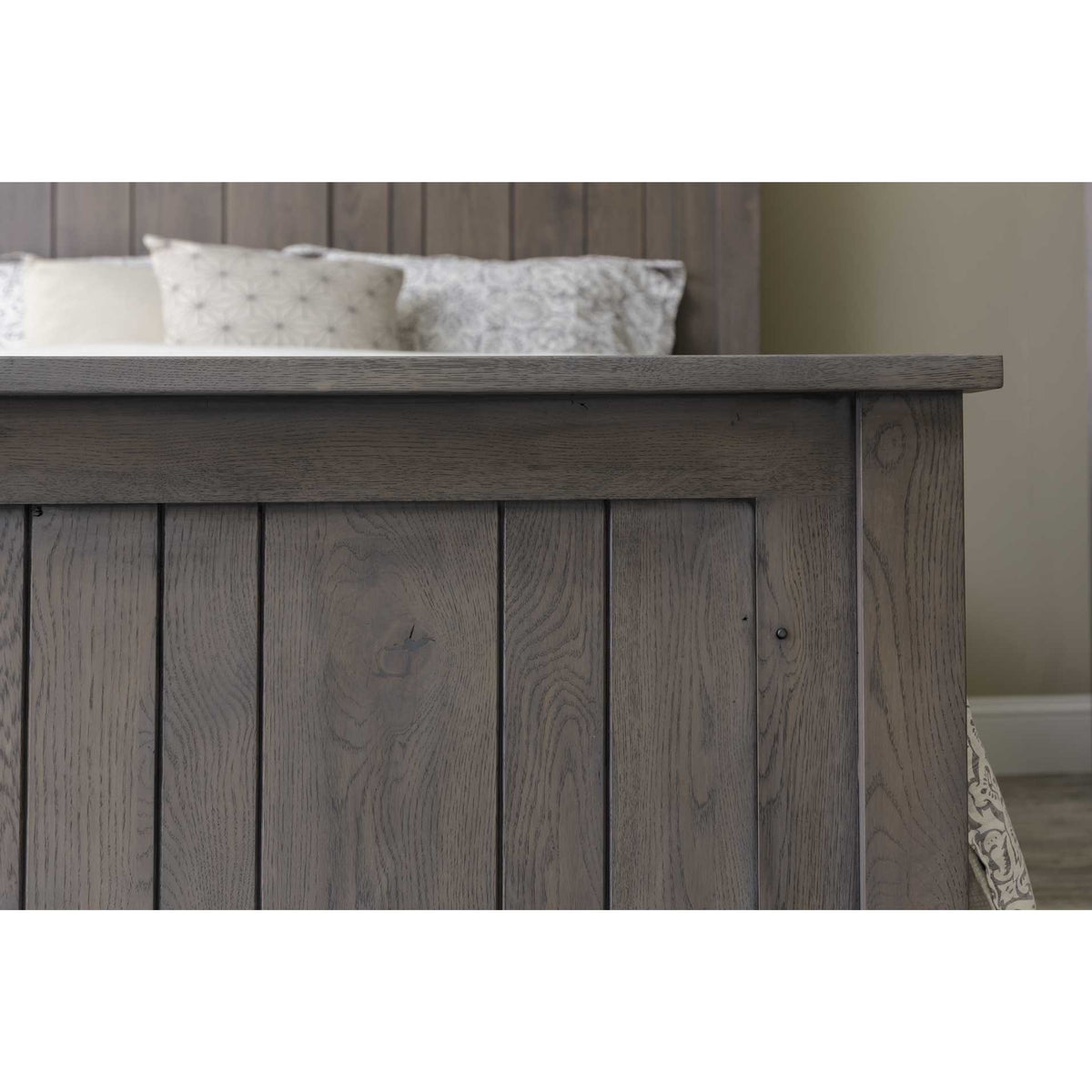 Corner of the gray plank footboard with its thick top rail, pillows and headboard visible beyond