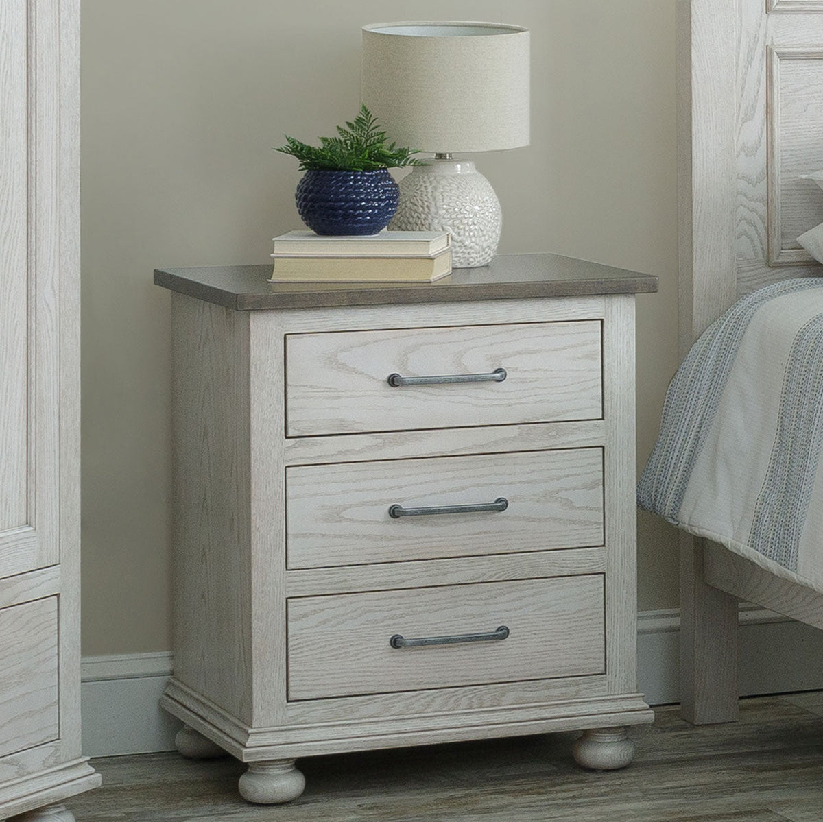Whitewashed three drawer nightstand with gray top, pewter bar pulls and bun feet, topped with a lamp and books