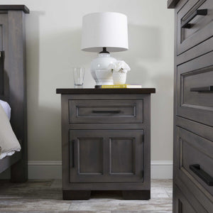 Gray solid wood nightstand with one drawer and a two-panel door, dark rustic top, styled with a white lamp between bed and dresser