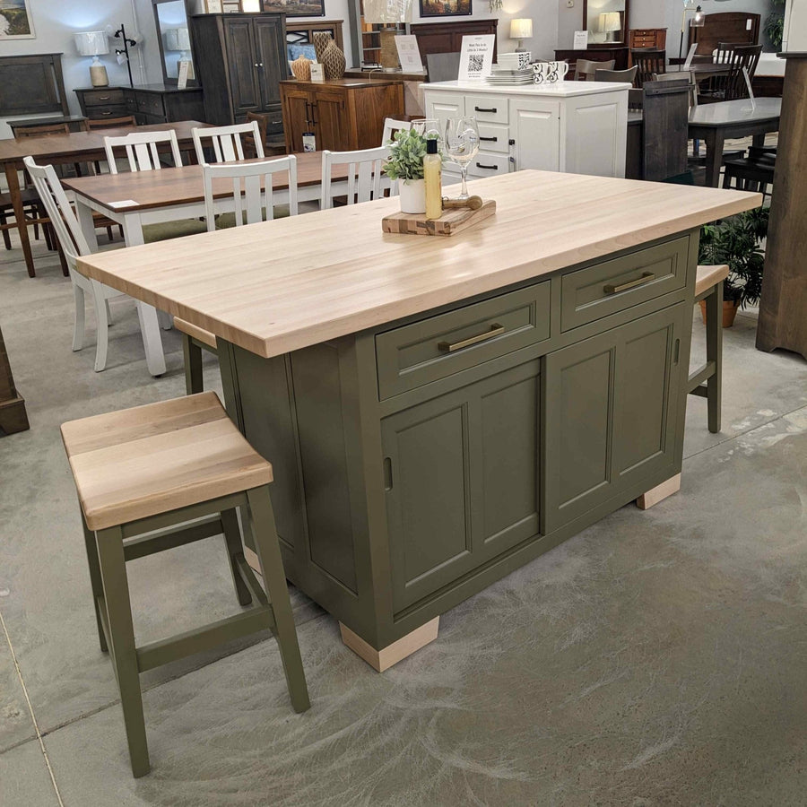 Sage green kitchen island with natural maple butcher block top and two matching saddle stools in the furniture showroom