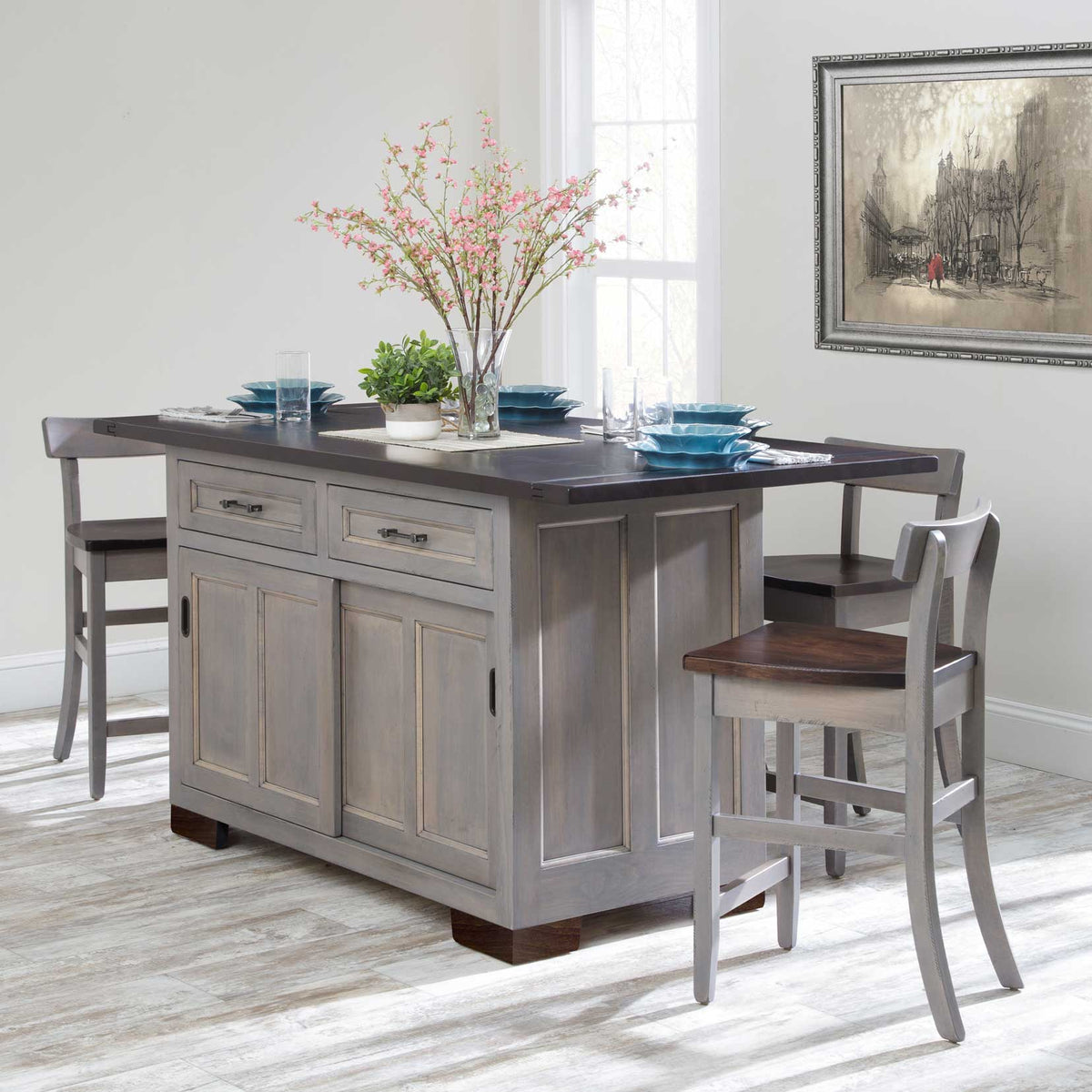 Gray kitchen island with dark stained overhang bar top, two drawers, sliding doors and matching counter stools set with blue dishes