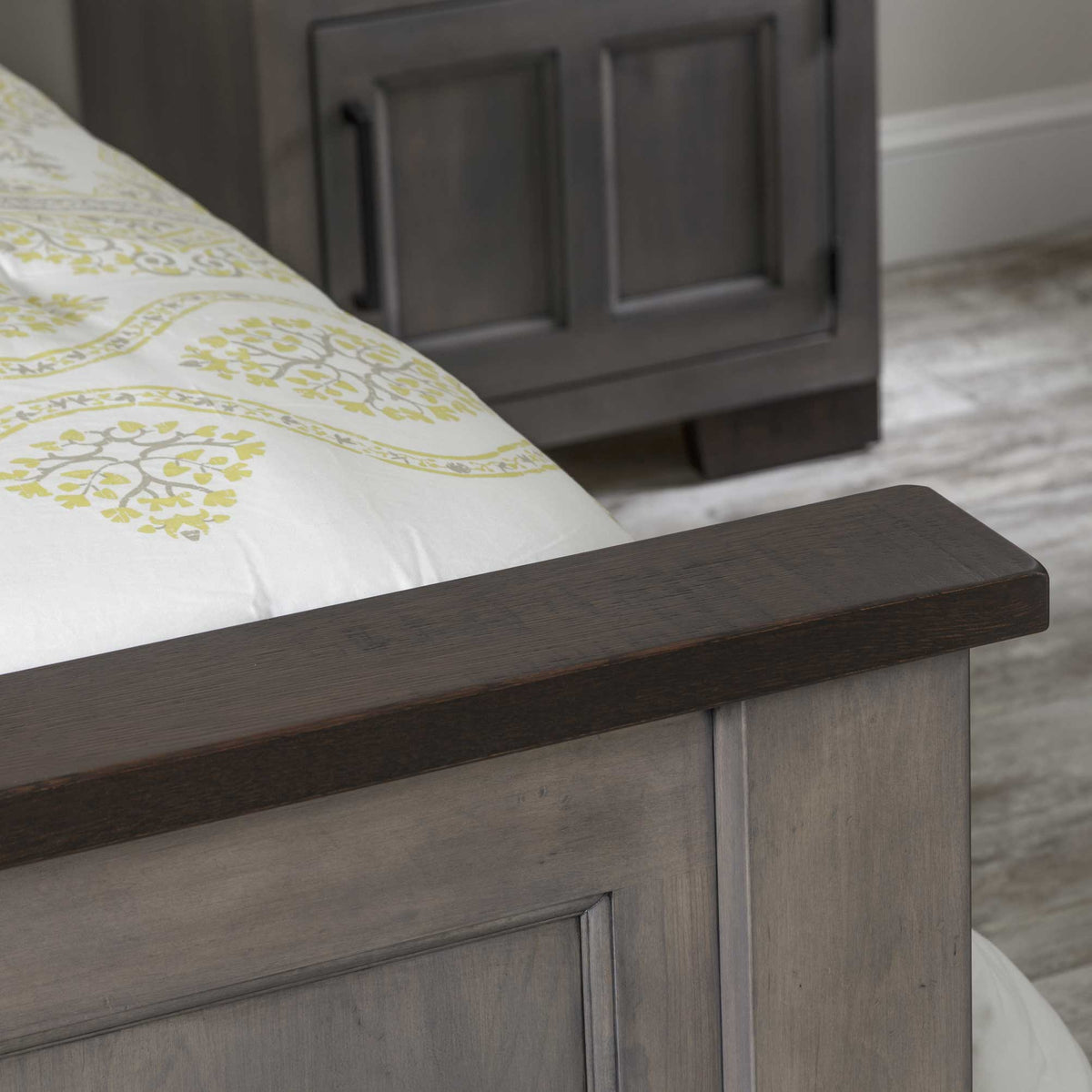 Close-up of the panel bed footboard corner in a gray two-tone finish showing the dark rustic top rail and matching nightstand behind