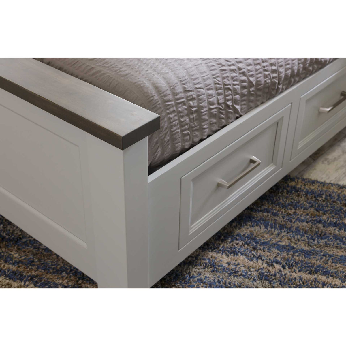 Footboard corner detail of the white storage bed showing the gray wood cap and raised-panel storage drawer with nickel bar pull