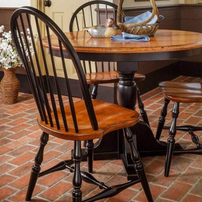 Round expandable pedestal dining table with cherry top and black base set with Windsor chairs in a brick floored kitchen
