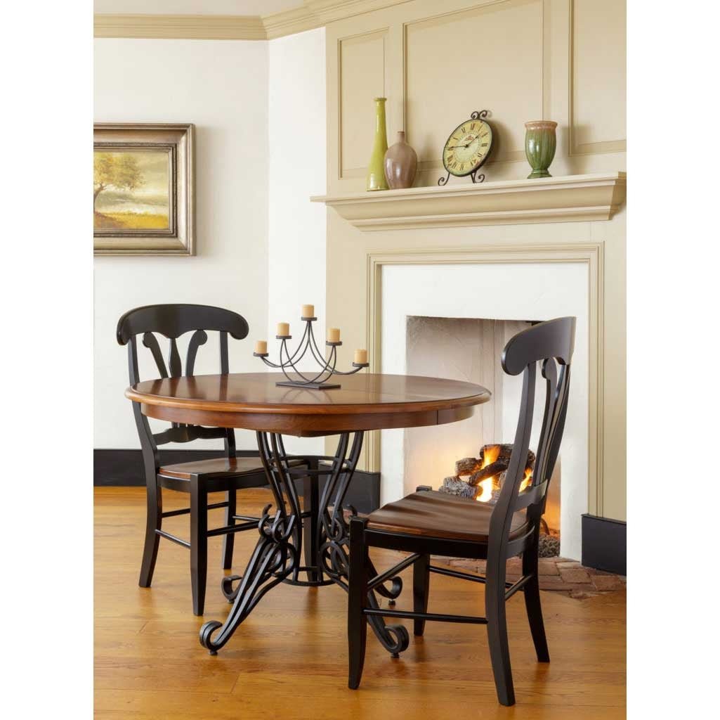 Round dining table with cherry top on a scrolled wrought iron base, flanked by two black chairs by a fireplace
