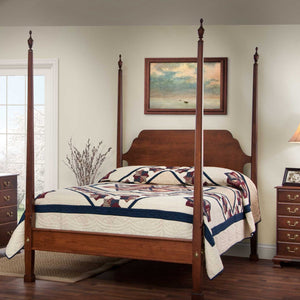 Cherry pencil post bed with four tall tapered posts, urn finials and a navy patchwork quilt in a sunny bedroom