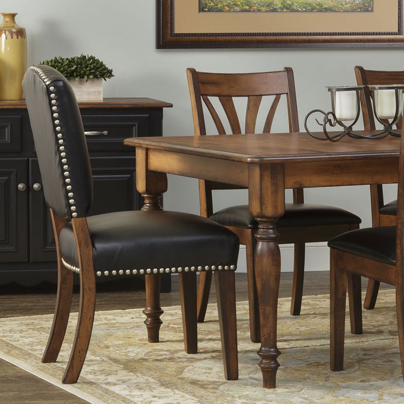 Close-up of a black leather upholstered dining chair with antique brass nailhead trim next to the turned-leg wood dining table