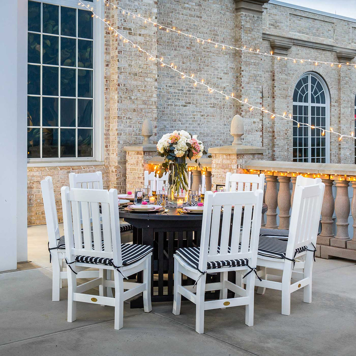 Round black poly patio dining table with six white slat back chairs and striped cushions under string lights