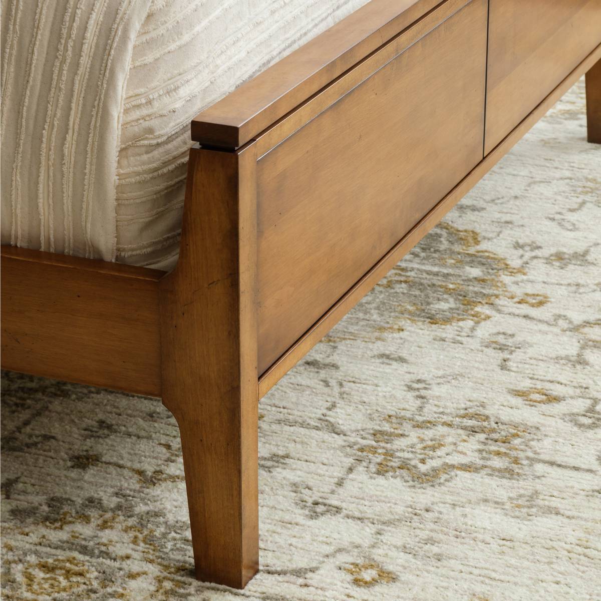 Low paneled footboard of the mid century maple bed with a stepped top rail and splayed leg on a patterned rug