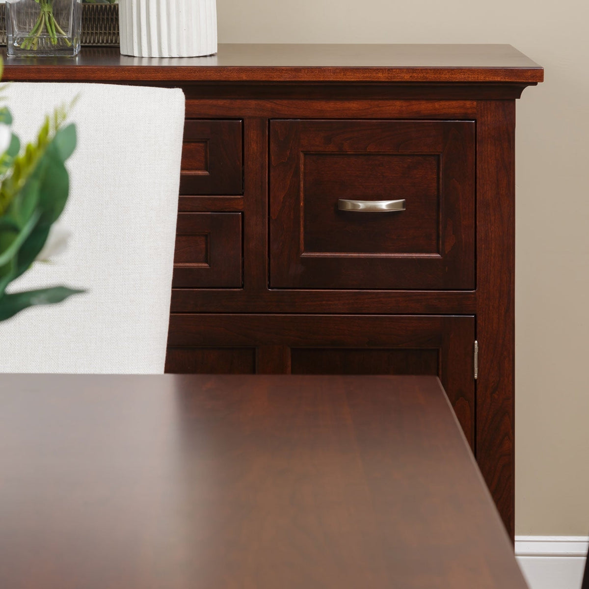 Corner detail of a cherry dining table top next to a buffet with inset drawers and brushed nickel pulls