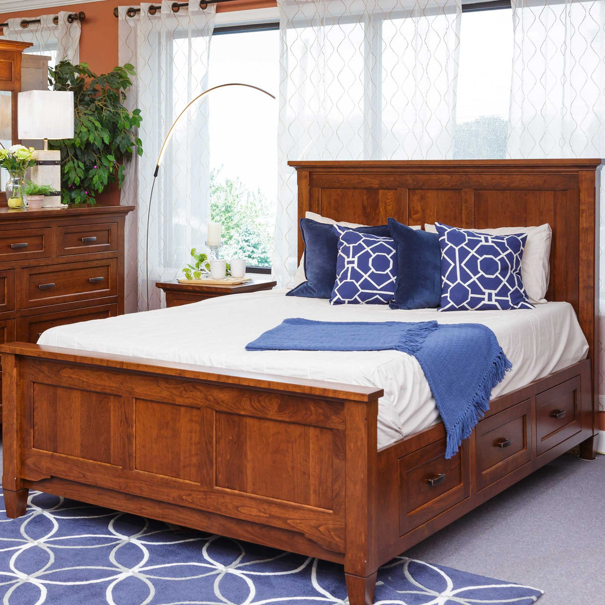 Storage bed in cherry with drawers at the foot, tall panel headboard and navy pillows in a sunlit bedroom