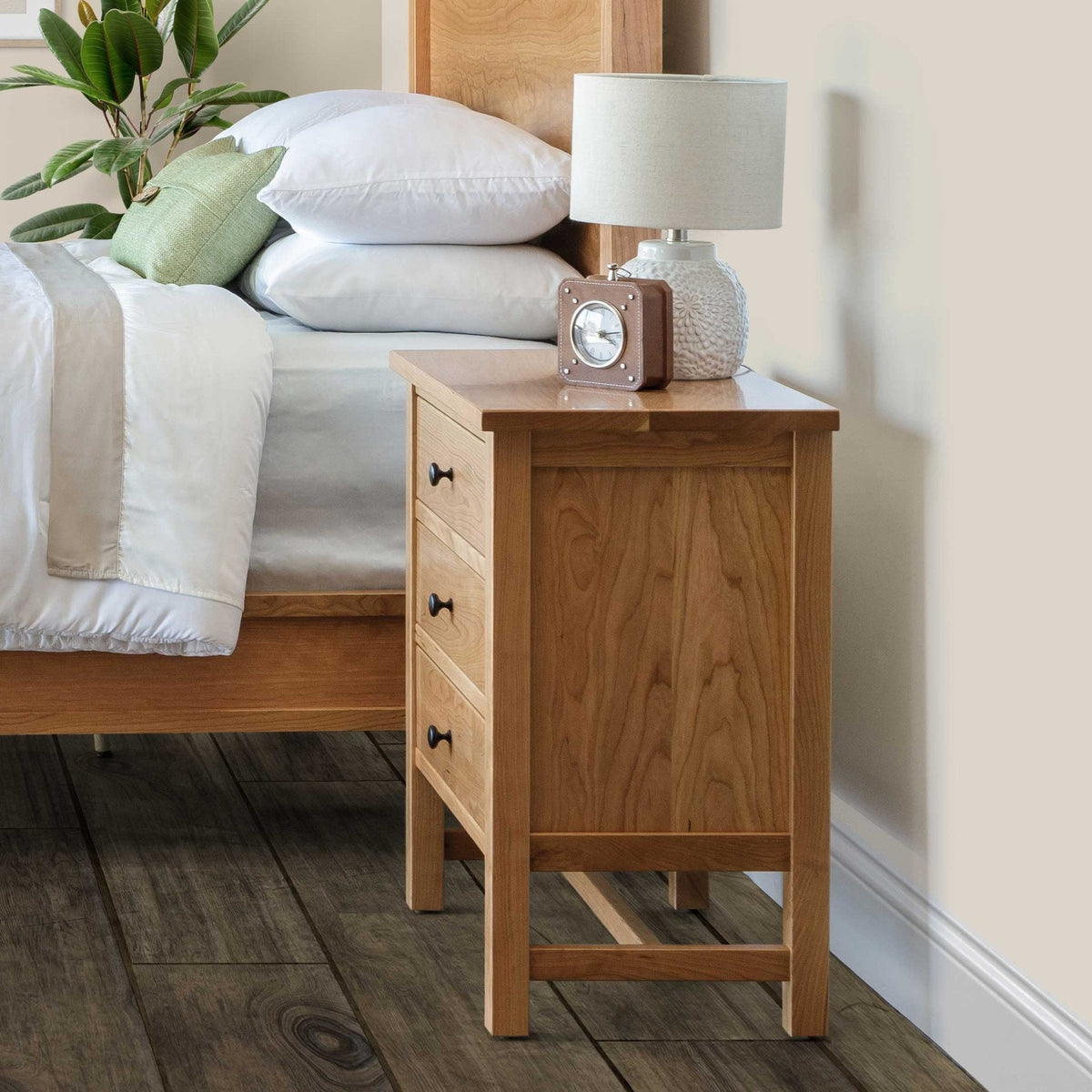 Side view of the natural cherry three-drawer nightstand beside a bed, topped with a ceramic lamp and small clock