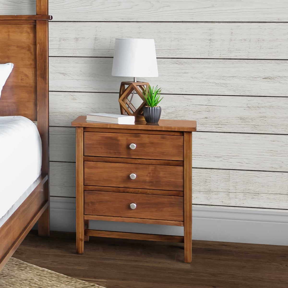 Three-drawer nightstand in a honey brown finish with nickel knobs styled beside a bed against white shiplap