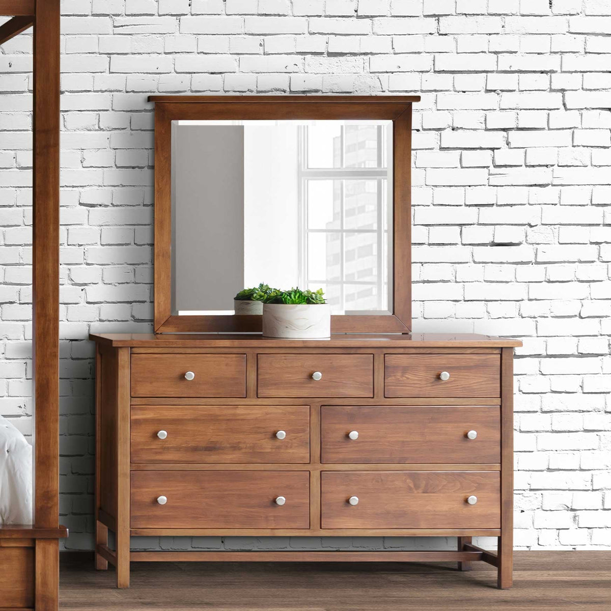 Seven-drawer dresser in a warm brown finish with white knobs and a square mirror holding potted succulents by a white brick wall