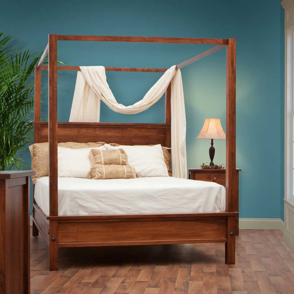 Canopy bed in a rich cherry finish with sheer draped fabric over the rails against a teal wall beside a lamp-lit nightstand