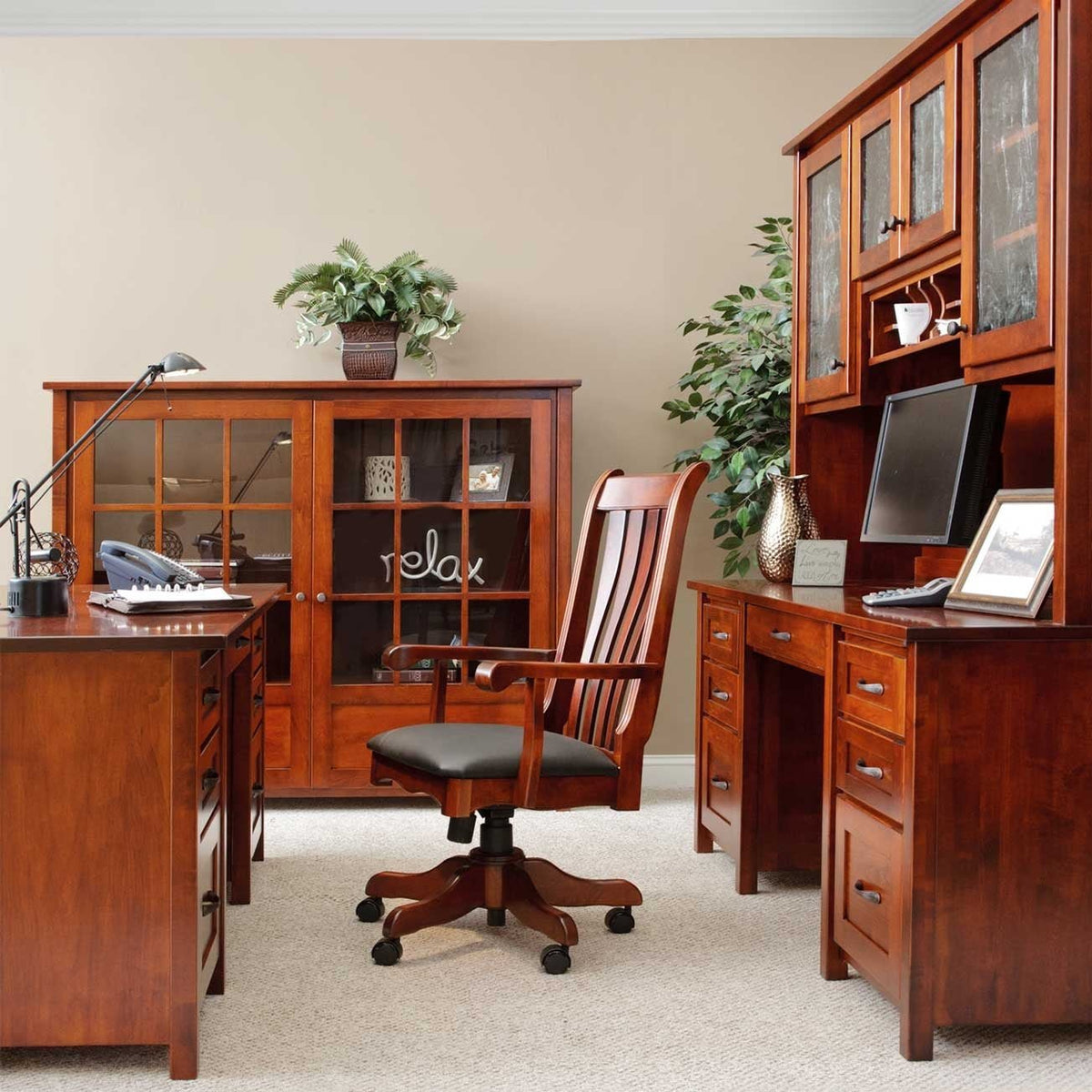 Home office set in cherry with an executive desk, credenza and hutch, glass-door bookcase and slat-back swivel chair on carpet
