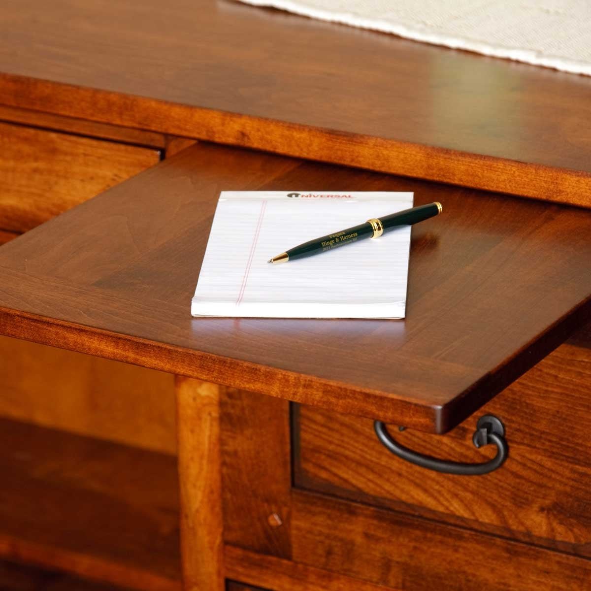 Sliding wood writing surface pulled out of a brown desk holding a lined notepad and green pen, black drawer pull below