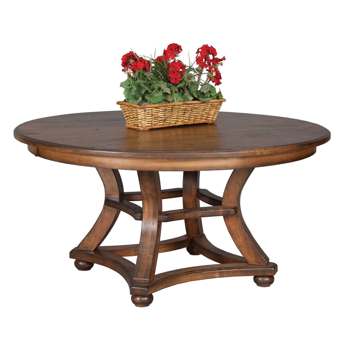Round dining table in a warm honey finish with a curved four-post base and a basket of red geraniums