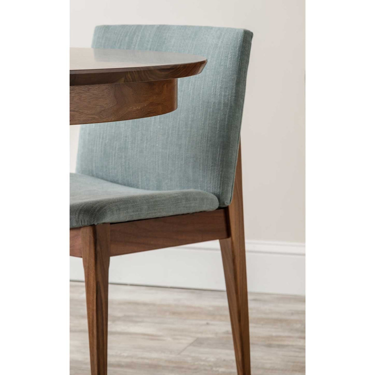 Side view of a green upholstered dining chair with an exposed walnut frame pulled up to a round table