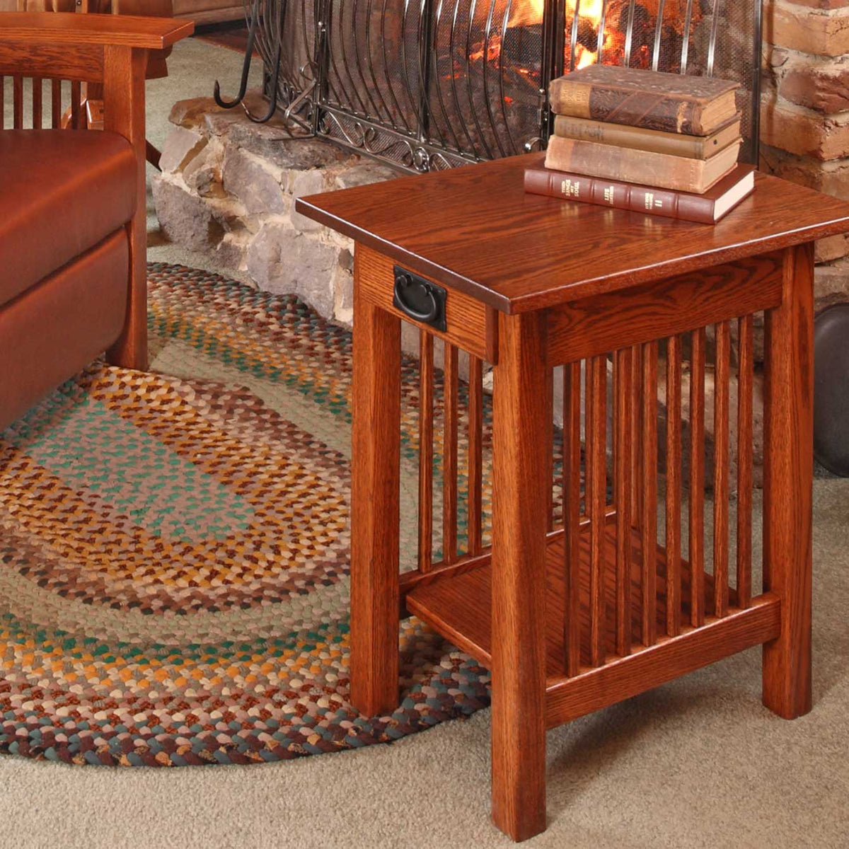 Oak chairside end table with drawer and slat sides holding stacked books, set on a braided rug by a stone fireplace