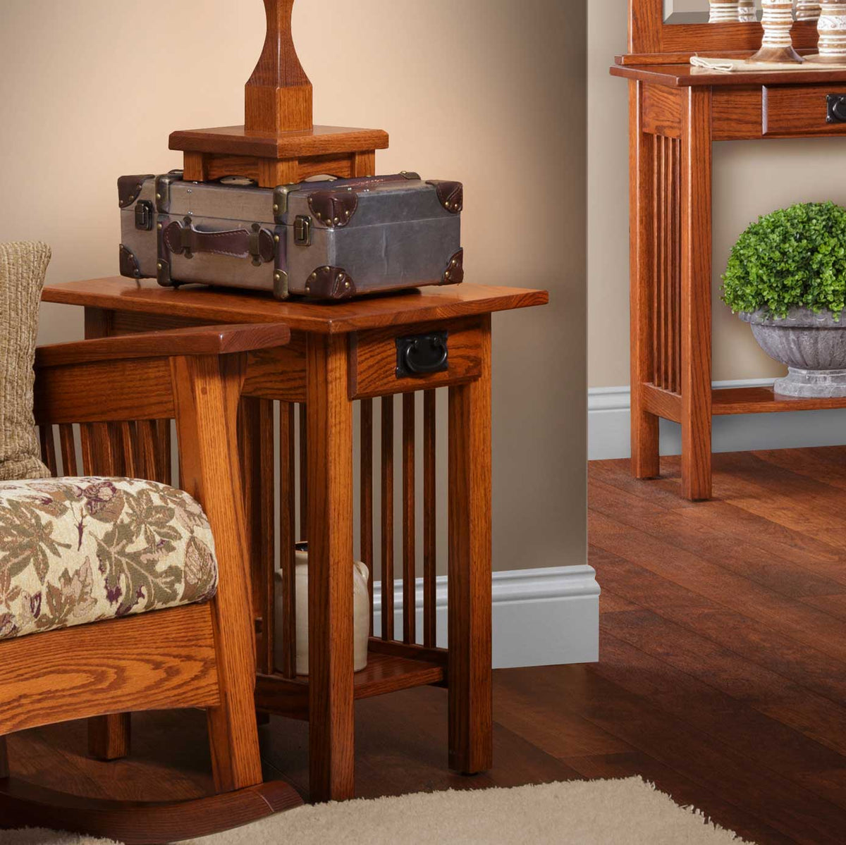 Mission oak end table with slatted sides and drawer holding a vintage suitcase and lamp beside an upholstered chair