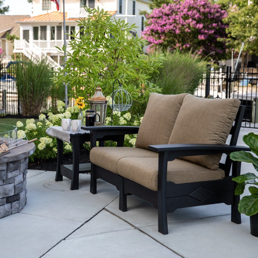 Black poly two seat patio loveseat with plush tan cushions and matching side table on a concrete patio by a garden