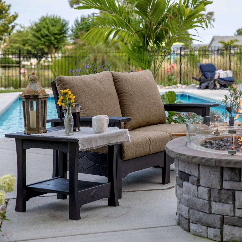 Black poly cushioned patio loveseat with tan cushions and a black end table beside a round stone gas fire pit