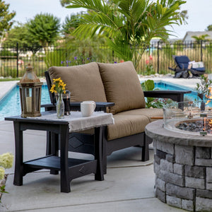 Black poly cushioned patio loveseat with tan cushions and a black end table beside a round stone gas fire pit
