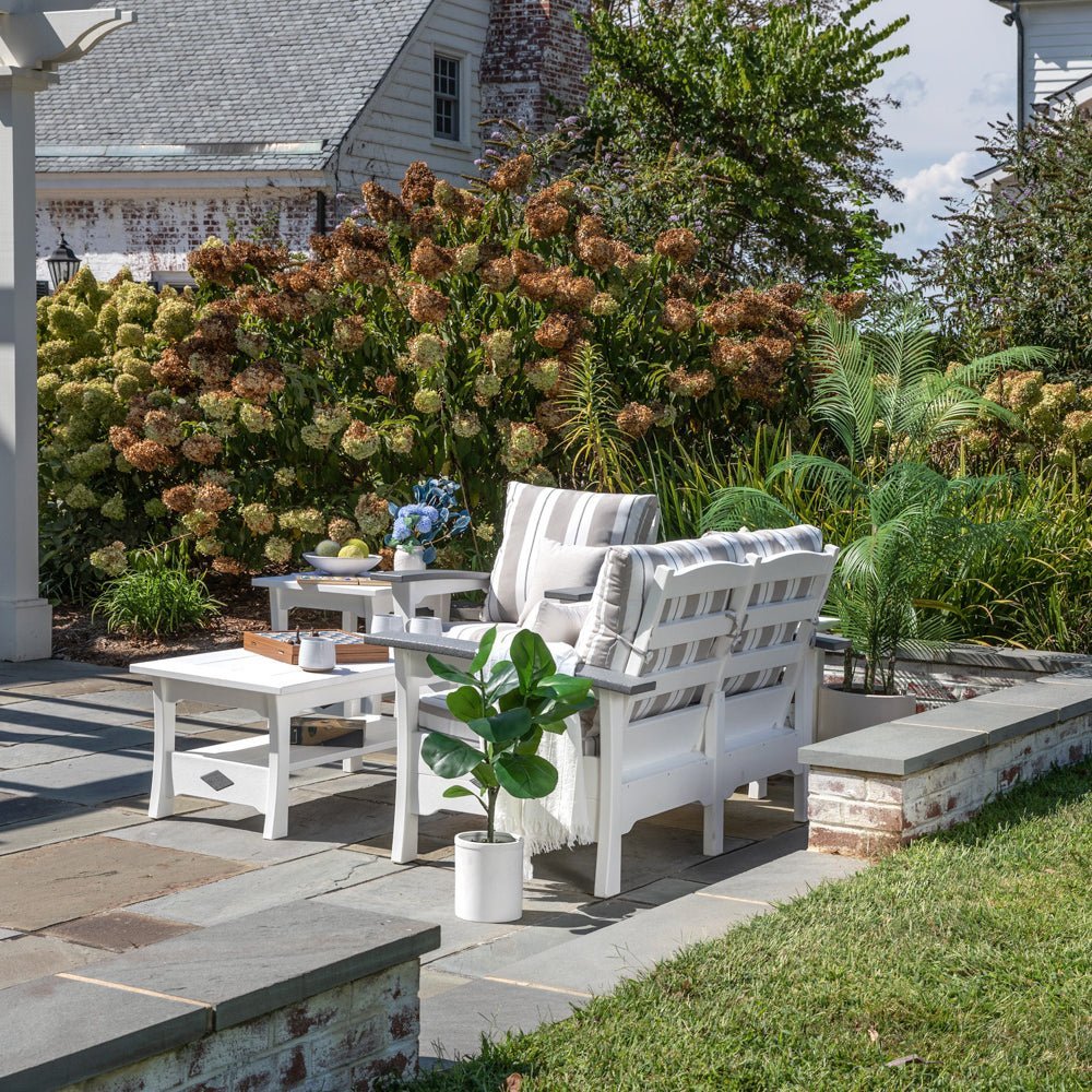 White poly loveseat set with gray striped cushions and two square end tables on a bluestone patio by hydrangeas